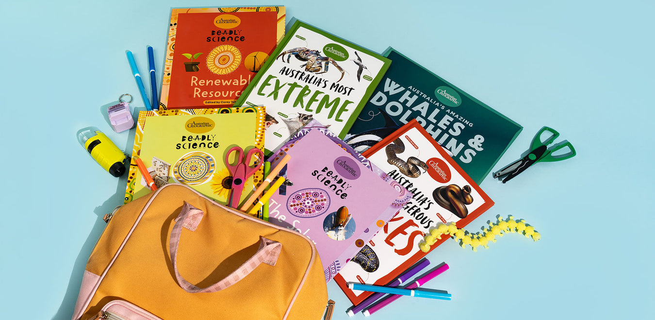 A curated flat-lay of children's activity and learning items arranged on a light blue background, including a mustard-yellow bag, colorful pens, scissors, a yellow sponge, and several books such as 'Renewable Resources' and 'Australian Animals'.