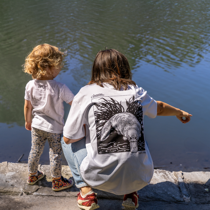 Two people, one adult and one child, standing by a body of water with a focus on the adult's shirt with a platypus design.
