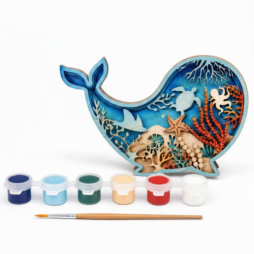 A whale-shaped wooden craft painting kit with an ocean-themed laser-cut design, six small paint pots, and a paintbrush on a white background.