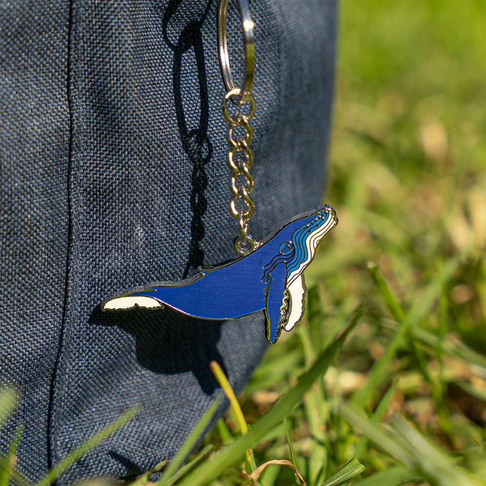 A blue and white whale-shaped keychain with a silver metal chain, shown hanging against a dark blue denim surface with blurred green grass in the background.