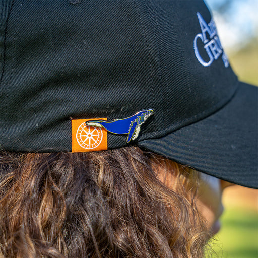 A close-up of a person wearing a black baseball cap outdoors, showing the back of the cap with a small orange square patch and a blue whale tail pin attached near the edge.