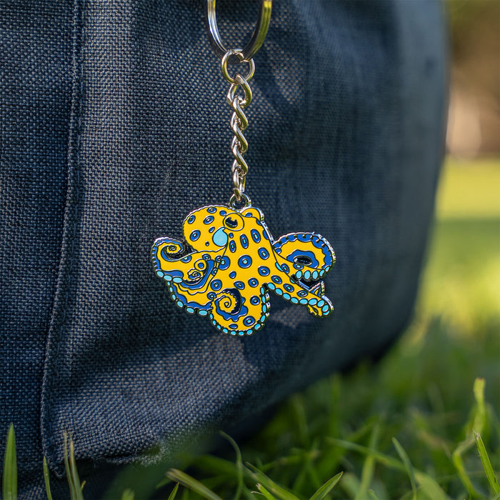 A yellow octopus-shaped keychain with blue and black spots and turquoise accents, hanging from a silver key ring against a dark denim bag with blurred green grass in the background.