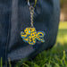 A yellow octopus-shaped keychain with blue and black spots and turquoise accents, hanging from a silver key ring against a dark denim bag with blurred green grass in the background.