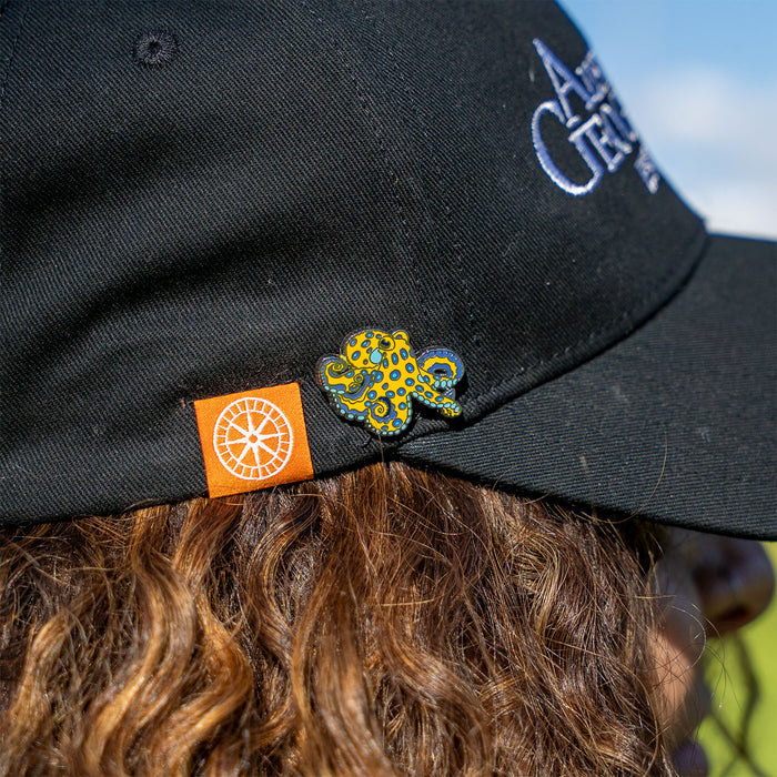 A close-up of a person wearing a black baseball cap outdoors. The cap has white embroidered text on the side and a small orange square patch near the brim with a white circular logo.