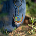 A colorful embroidered keychain featuring a stylized spider design with a yellow and blue patterned body, red details, and silver-toned metal hardware, shown hanging against a blue denim-clad person outdoors with blurred greenery in the background.