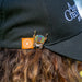 A close-up lifestyle shot of a person wearing a black baseball cap with embroidered white text, brown hair, and two small enamel pins clipped to the brim. The pins include an orange square with a white geometric design and a silver-toned shield-shaped pin with colorful details.