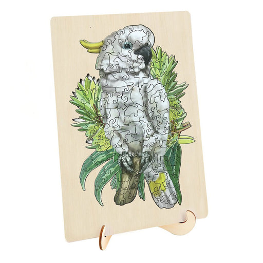 A rectangular wooden jigsaw puzzle board featuring an illustrated white cockatoo perched among green leaves, displayed on a small wooden stand against a plain white background.