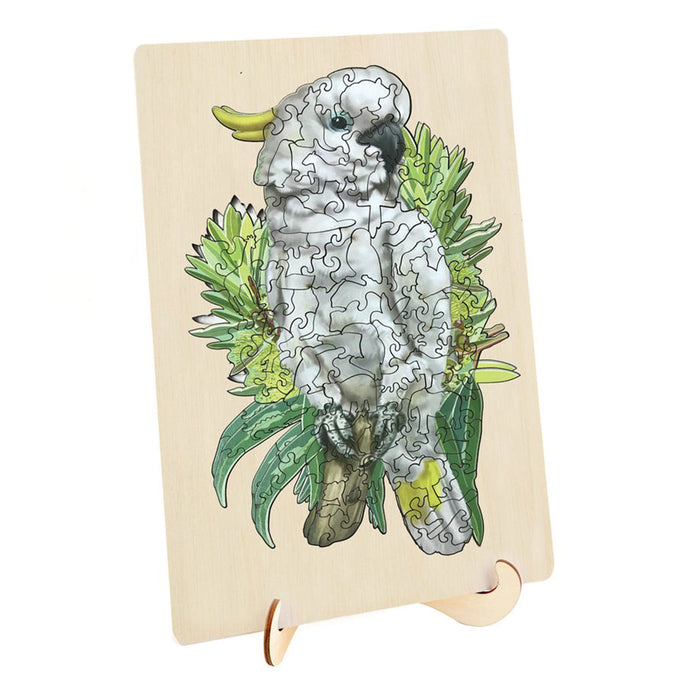 A rectangular wooden jigsaw puzzle board featuring an illustrated white cockatoo perched among green leaves, displayed on a small wooden stand against a plain white background.