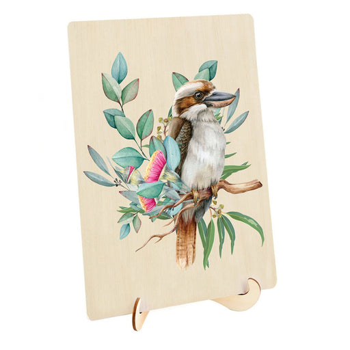 A rectangular wooden plaque or sign on a small wooden display stand, featuring a painted kookaburra perched among green leaves and pink flowers on a light wood background.