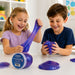 Two children, a girl in a lavender shirt stretching a purple slime and a boy in a navy shirt holding a purple slime ball, sitting at a white table with a jar of Starbrightz Glittery Slime.