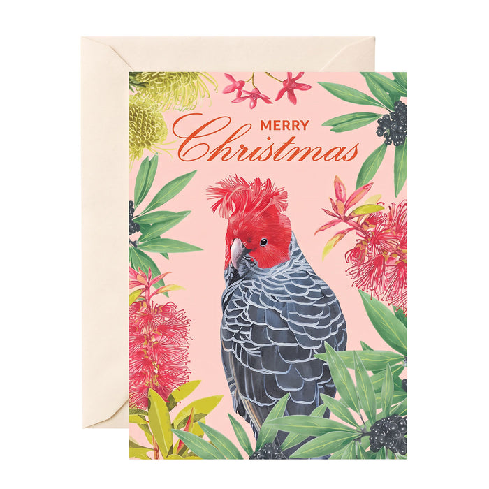 A Christmas greeting card with a pink background, featuring a red cockatoo surrounded by tropical flowers and green leaves, with the words 'Merry Christmas' at the top. The card is shown with a cream-colored envelope behind it on a white background.