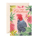 A Christmas greeting card with a pink background, featuring a red cockatoo surrounded by tropical flowers and green leaves, with the words 'Merry Christmas' at the top. The card is shown with a cream-colored envelope behind it on a white background.