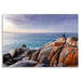 A horizontal landscape art print depicting a cyclist on a rocky, orange-lichenated coast with turquoise ocean water under a blue and purple sky. Small text is visible in the upper left corner.