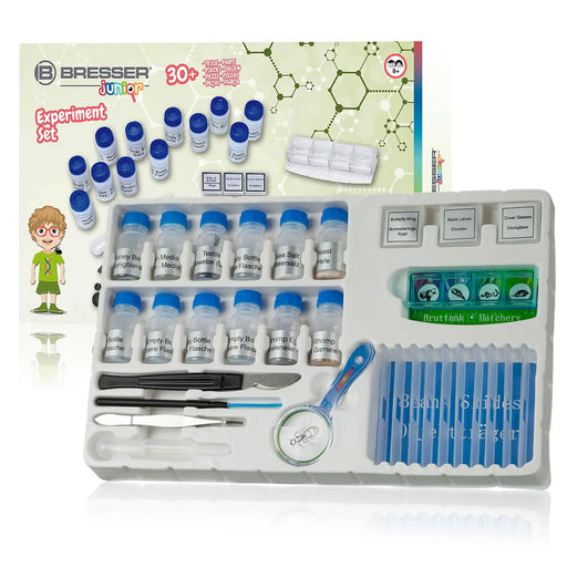 Bresser Junior Chemistry Experiment Set in a white plastic tray, featuring 30+ small vials with blue caps, test tubes, tweezers, a scalpel, a magnifying glass, and a 4-color indicator strip, shown alongside its illustrated packaging box.