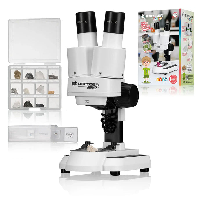 Bresser Biolux 2X educational toy microscope set, featuring a white and black binocular microscope, a clear specimen storage box filled with small rocks, a slide tray, and a colorful retail packaging box in the background.
