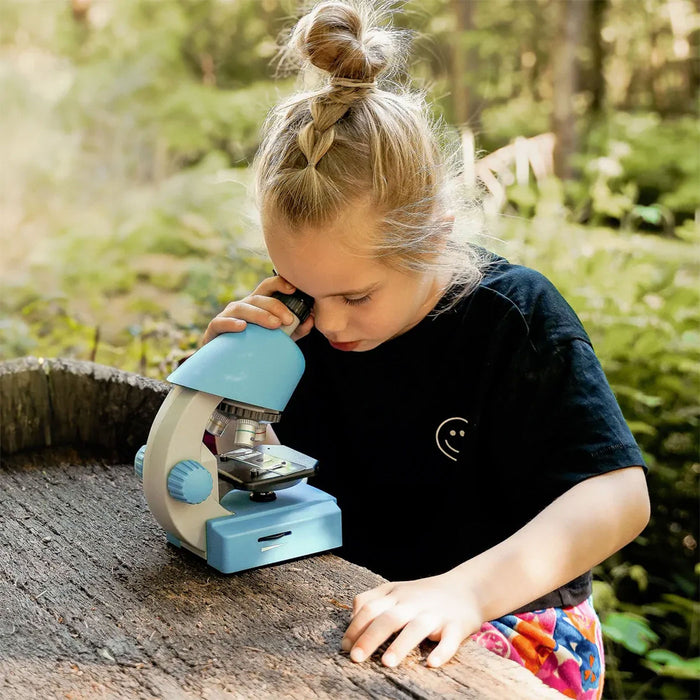 A young girl with blonde hair in a bun looks through a light blue and white toy microscope placed on a wooden surface outdoors in a sunlit forest setting.