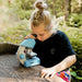 A young girl with blonde hair in a bun looks through a light blue and white toy microscope placed on a wooden surface outdoors in a sunlit forest setting.