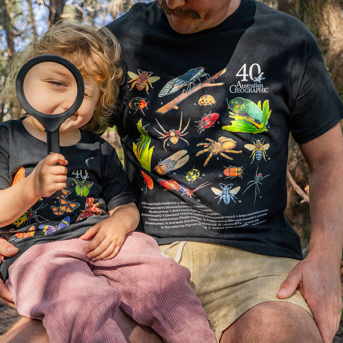 A man and a young child wearing matching black graphic t-shirts outdoors. The shirts feature colorful illustrations of insects, spiders, and other bugs, with the text '40 Years Australian Geographic' on the man's shirt.