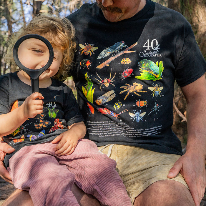 A man and a young child wearing matching black graphic t-shirts outdoors. The shirts feature colorful illustrations of insects, spiders, and other bugs, with the text '40 Years Australian Geographic' on the man's shirt.