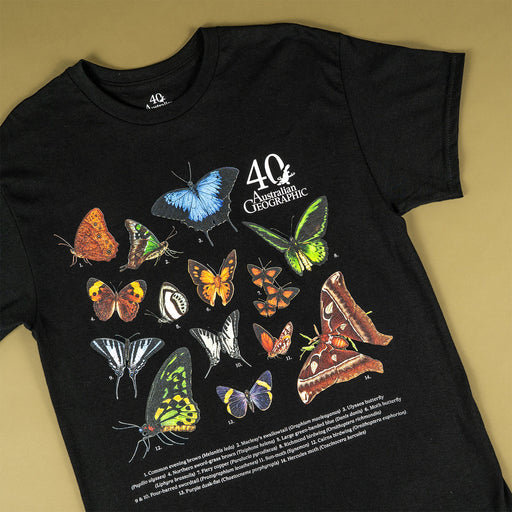 A black short-sleeve T-shirt laid flat on a warm beige background, featuring a large printed collage of colorful butterflies and moths with the text '40 Australian Geographic' and small caption text near the bottom.