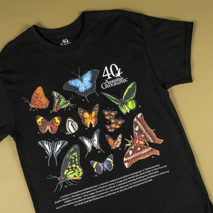 A black short-sleeve T-shirt laid flat on a warm beige background, featuring a large printed collage of colorful butterflies and moths with the text '40 Australian Geographic' and small caption text near the bottom.