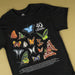A black short-sleeve T-shirt laid flat on a warm beige background, featuring a large printed collage of colorful butterflies and moths with the text '40 Australian Geographic' and small caption text near the bottom.
