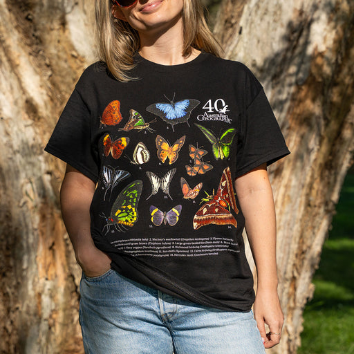 A woman wearing a black graphic t-shirt with a colorful butterfly print and '40th Anniversary' text, standing outdoors in front of a tree trunk.