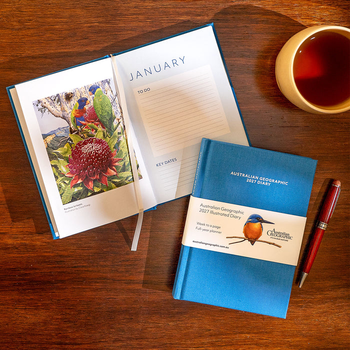 An open and closed blue Australian Geographic 2021 Recommended Diary are arranged on a wooden table, alongside a cup of tea, a red pen, and a bird illustration printed inside the open book.