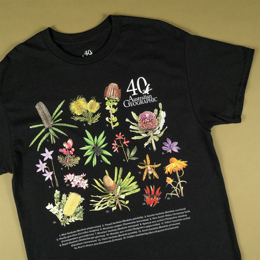 A black short-sleeve graphic T-shirt laid flat on a warm olive-beige background. The shirt features a large, colorful botanical illustration of Australian flora with the text '40 Australian Geographic' and small caption text at the bottom.