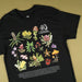 A black short-sleeve graphic T-shirt laid flat on a warm olive-beige background. The shirt features a large, colorful botanical illustration of Australian flora with the text '40 Australian Geographic' and small caption text at the bottom.