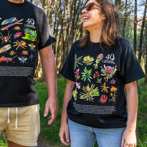 Two people standing outdoors in a wooded area wearing matching black graphic T-shirts. The shirts feature colorful illustrations of insects, plants, and the text '40 Australian Geographic.