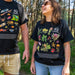 Two people standing outdoors in a wooded area wearing matching black graphic T-shirts. The shirts feature colorful illustrations of insects, plants, and the text '40 Australian Geographic.