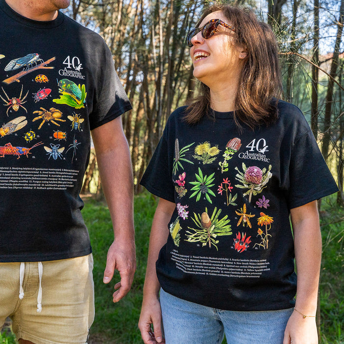 Two people standing outdoors in a wooded area wearing matching black graphic T-shirts. The shirts feature colorful illustrations of insects, plants, and the text '40 Australian Geographic.