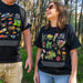 Two people standing outdoors in a wooded area wearing matching black graphic T-shirts. The shirts feature colorful illustrations of insects, plants, and the text '40 Australian Geographic.