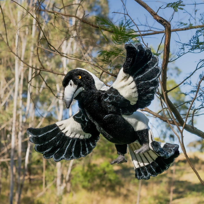 Plush toy magpie in flight with a natural background of trees and sky.