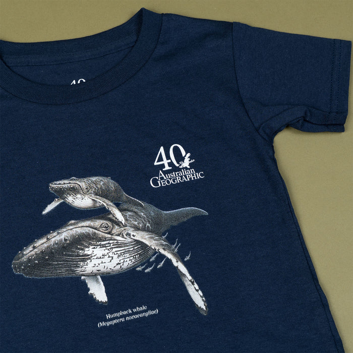 A navy blue short-sleeve t-shirt featuring a white graphic print of two humpback whales and the text '40 Australian Geographic' on the chest, laid flat on a solid olive green background.