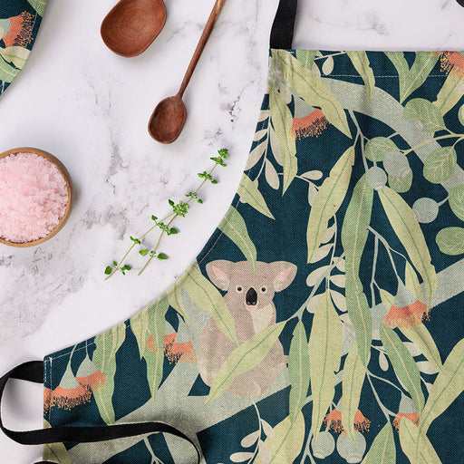 Apron with koala and eucalyptus design on a marble surface with kitchen items.