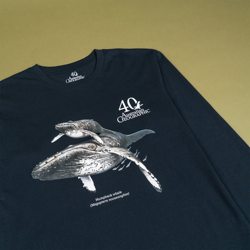 A dark navy long-sleeve t-shirt laid flat on a solid olive-green background. The shirt features a detailed graphic print of two humpback whales in white and gray, with '40th Australian Geographic ' and 'Humpback Whale (Megaptera novaeangliae)' printed in white text.