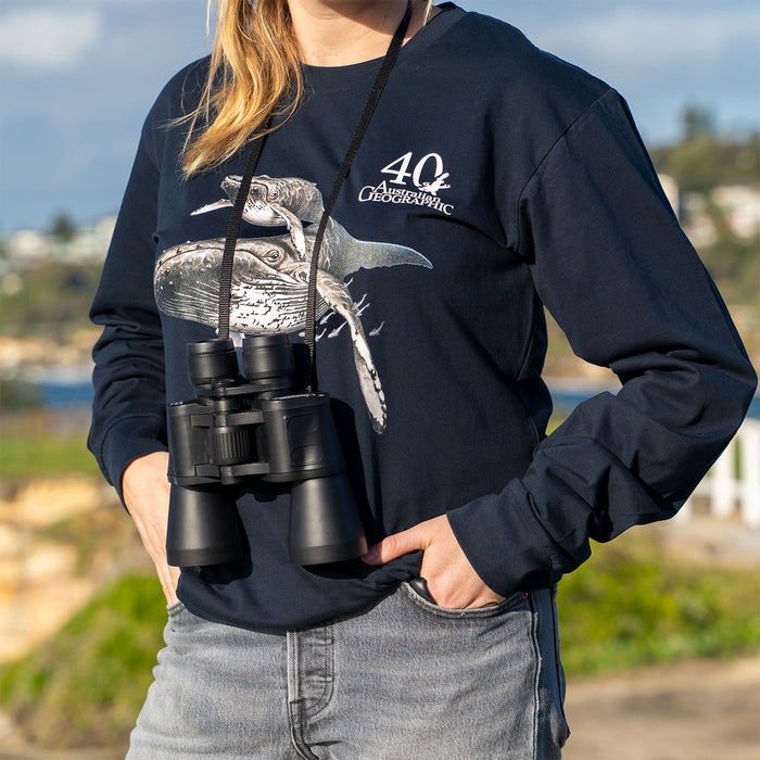 A person outdoors wearing a navy blue long-sleeve graphic sweatshirt with a whale print and '40 Australian Geographic' text, holding black binoculars around the neck. The background shows a blurred beach and sky.