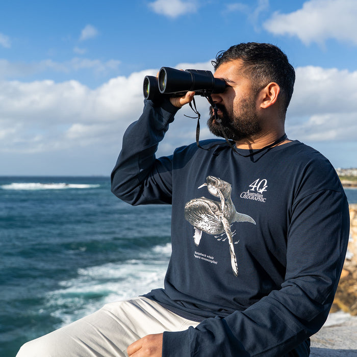 A man with a beard wearing a navy blue long-sleeve t-shirt with a sea turtle graphic and the text '40 Australian Geographic' looks through binoculars with an ocean and cloudy sky in the background.