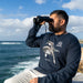 A man with a beard wearing a navy blue long-sleeve t-shirt with a sea turtle graphic and the text '40 Australian Geographic' looks through binoculars with an ocean and cloudy sky in the background.