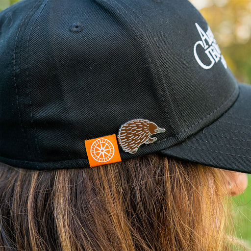 Black cap with a logo and echidna pin, worn by a person outdoors.