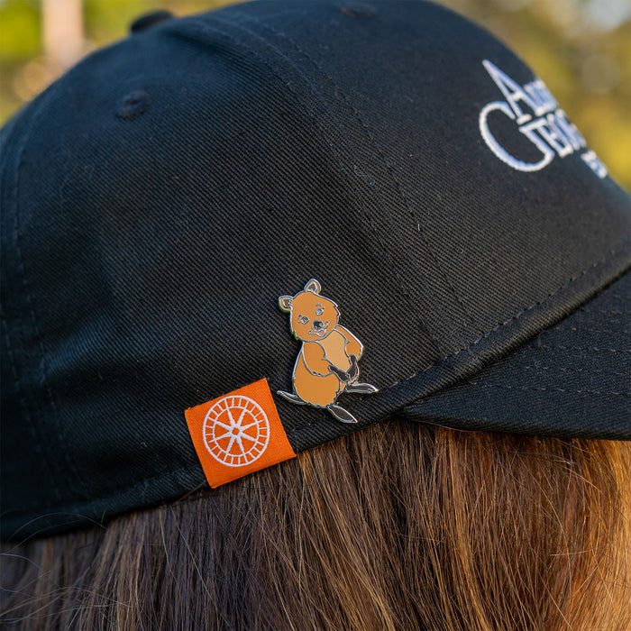 Black cap with a small quokka pin and orange label worn by a person with brown hair.