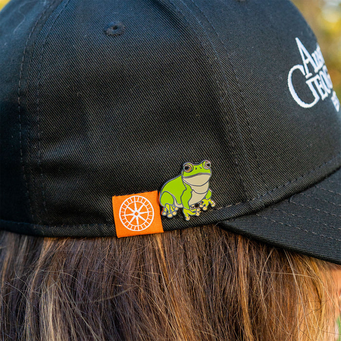 Black cap with a green frog pin and orange tag worn by a person