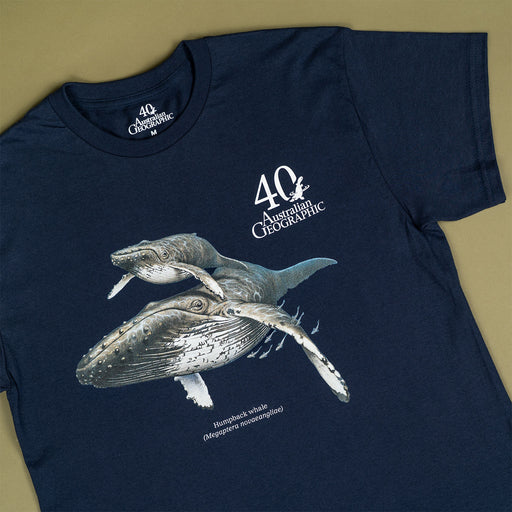 A navy blue short-sleeve t-shirt featuring a graphic print of two humpback whales and the text '40th Australian Geographic' on the front, laid flat on a beige background.