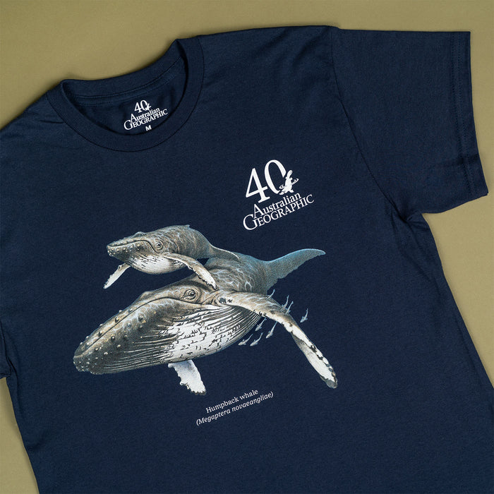 A navy blue short-sleeve t-shirt featuring a graphic print of two humpback whales and the text '40th Australian Geographic' on the front, laid flat on a beige background.