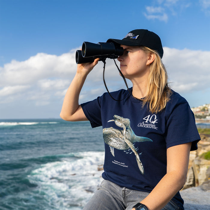 A woman wearing a navy blue t-shirt with a whale graphic and '40 Australian Geographic' text, along with a black cap, looking through binoculars by the ocean.
