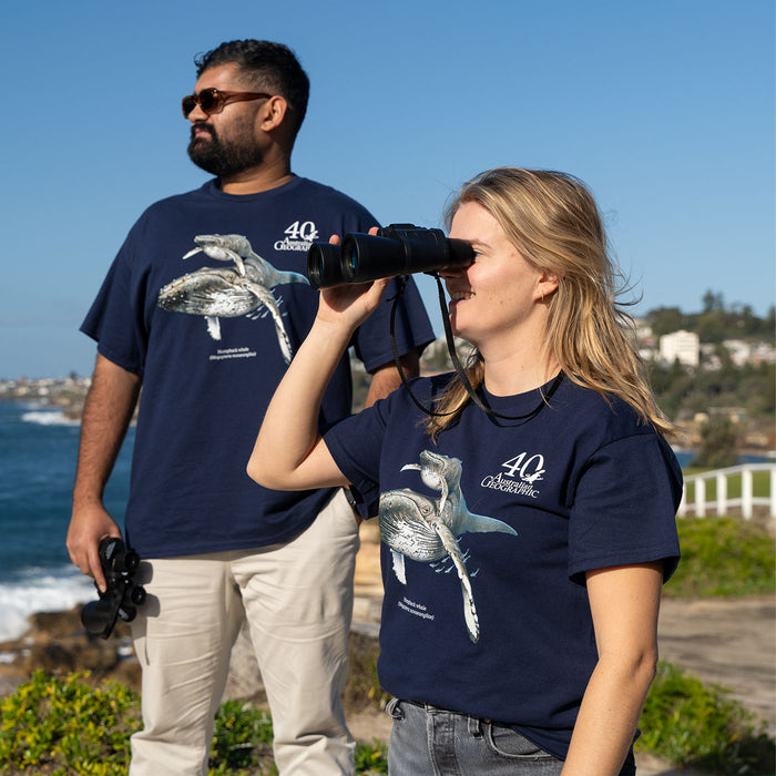 A man and a woman standing outdoors by the ocean, both wearing matching navy blue t-shirts with a whale graphic and the text '40 Australian Geographic'. The woman is looking through binoculars, and the man is holding binoculars in his hand.