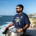 A man wearing a navy blue graphic t-shirt with a whale print sits on a rocky cliff overlooking the ocean, holding a magazine and binoculars.