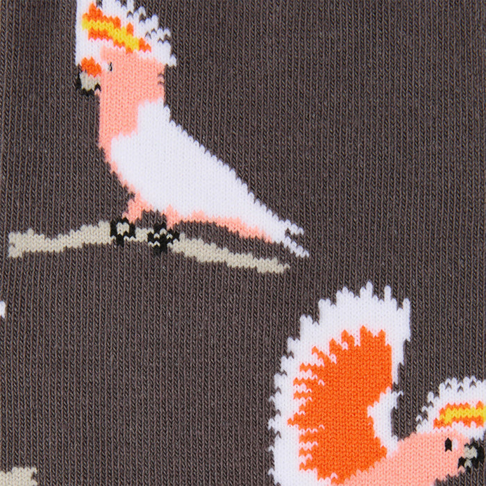 Close-up of a knit fabric featuring a repeating pattern of pink and white cockatoos perched on branches against a dark brown background.